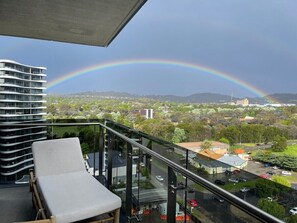 Een balkon met glazen hekwerk met uitzicht op een stadsgezicht, een witte lounge stoel en een regenboog op de achtergrond.