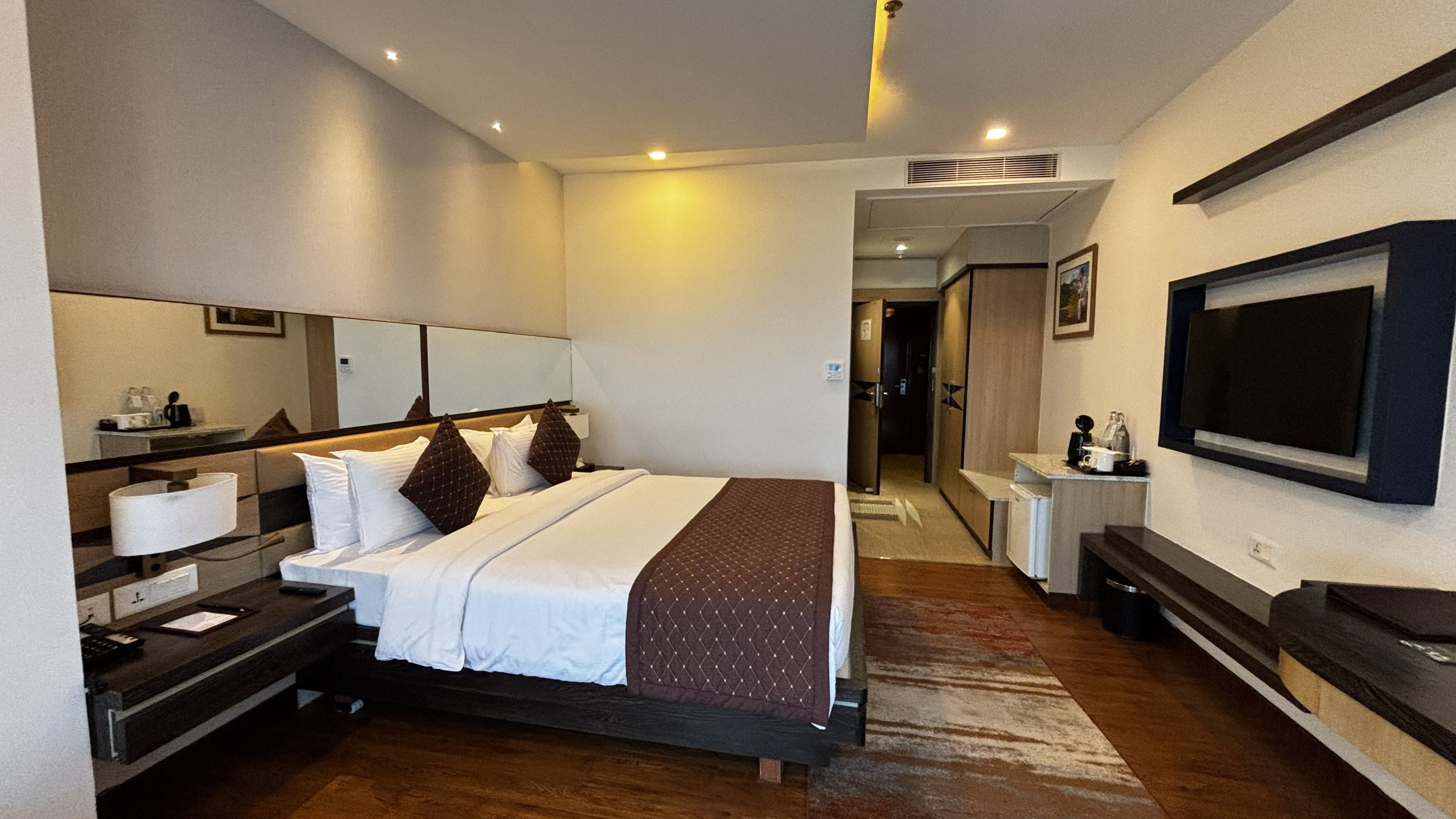 Quarto de hotel com cama, mesas de cabeceira, televisão, espelho e banheiro visível por uma porta aberta.