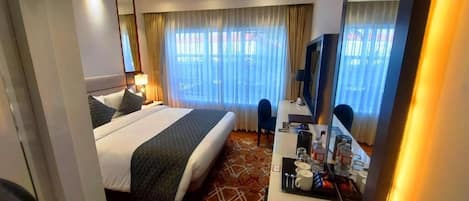 A hotel room with a bed, a desk with a chair, a TV, and a window with curtains.