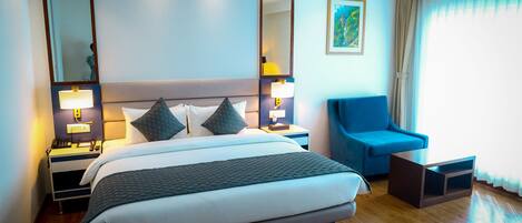 Una habitación de hotel moderna con una cama grande, un sillón azul, una mesita de noche con lámparas y un espejo amplio.