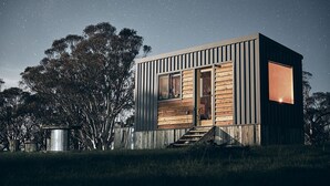 Una cabaña moderna y compacta con fachada de madera y ventanas grandes, rodeada de árboles bajo un cielo estrellado.
