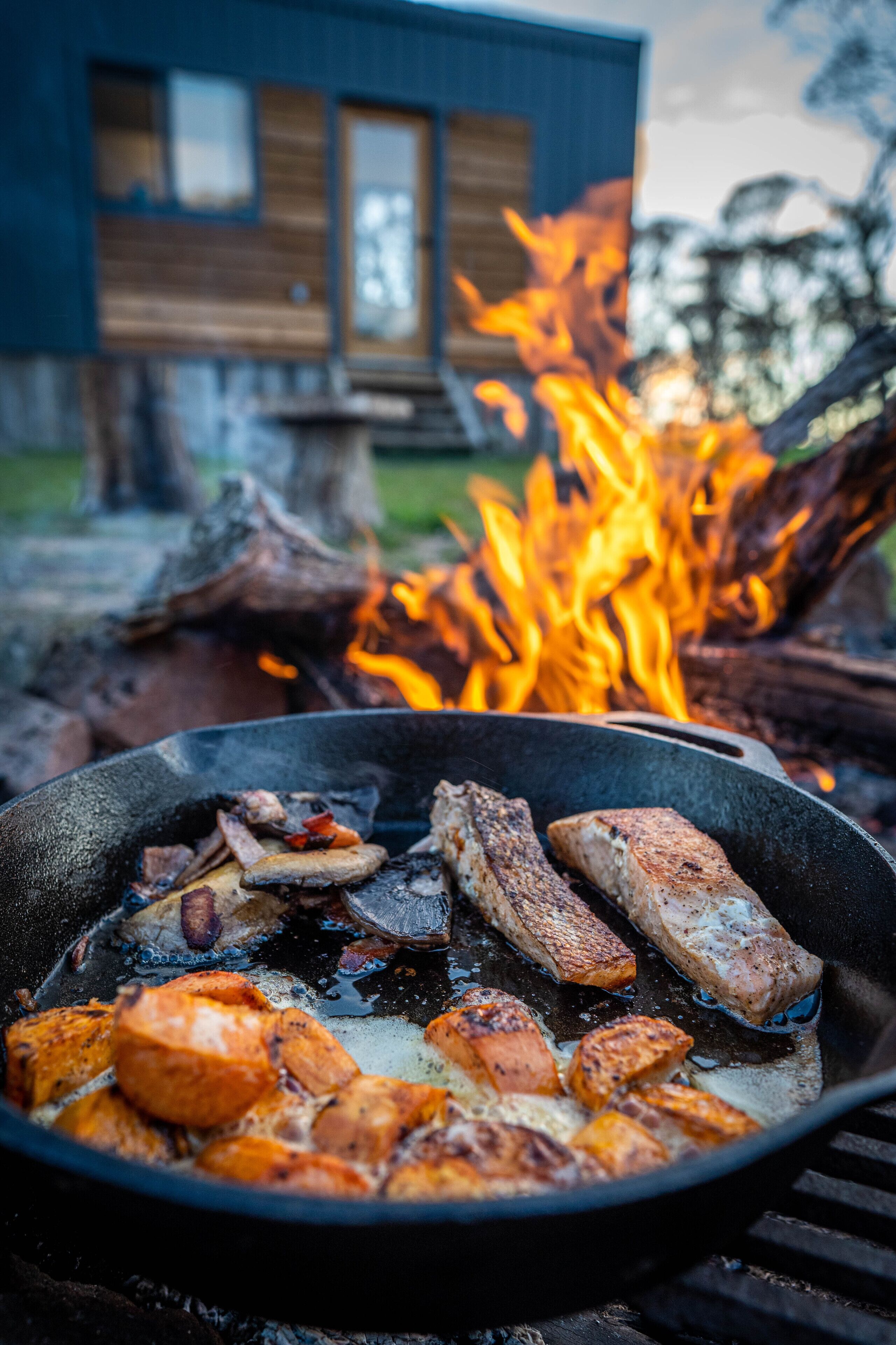 A skillet with grilled meat and vegetables over an open fire, with a modern building in the background.