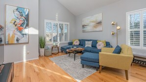 A modern living room with a blue sofa, a yellow armchair, and a coffee table. There are two large windows with blinds, a floor lamp, and a painting on the wall. - Super Cute Top Location, Fast WiFi (Reno)