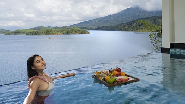 Una mujer en una piscina con una bandeja de frutas y pan, con vistas a un lago tranquilo y montañas.
