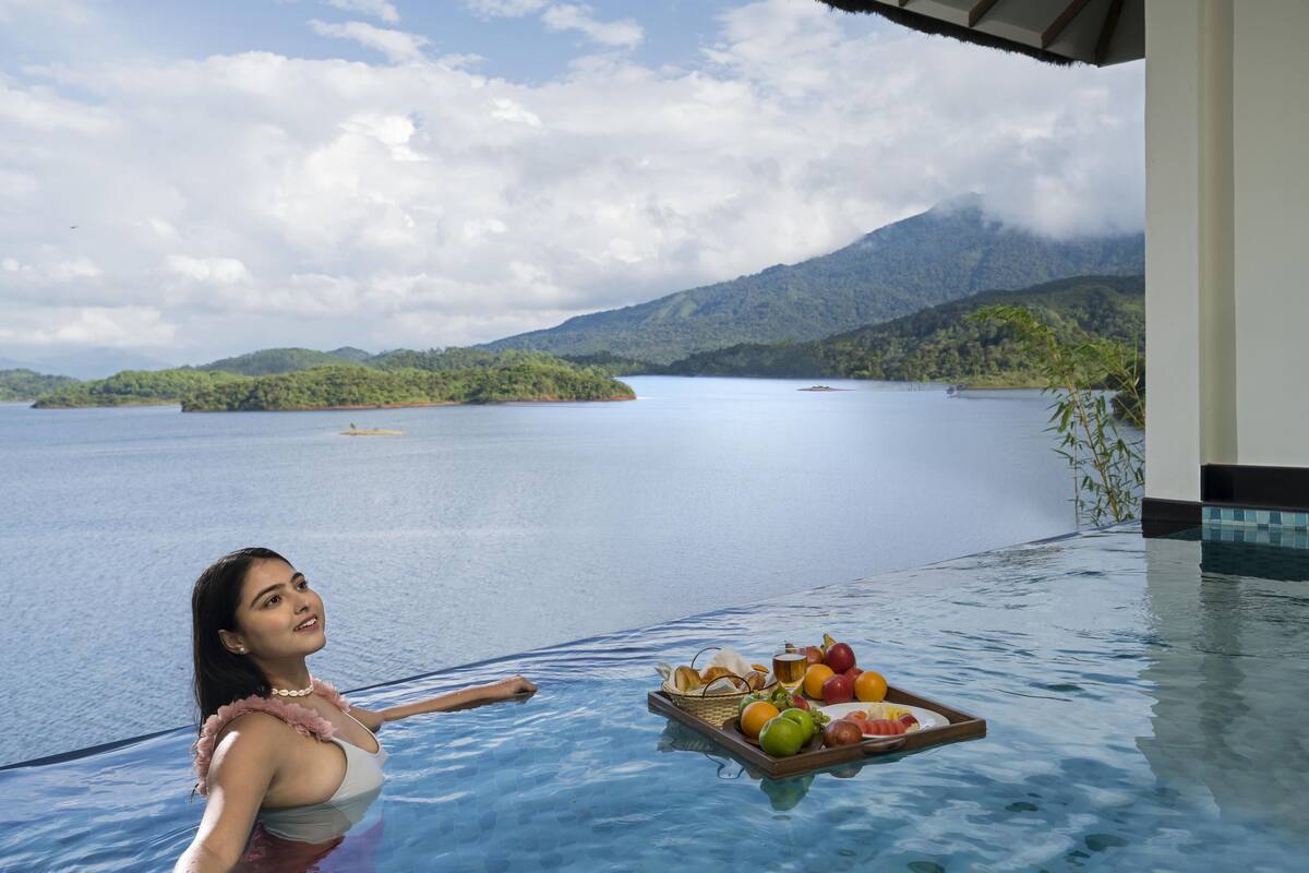 Mulher em piscina com bandeja de frutas e pão, com vista para um lago calmo e montanhas.