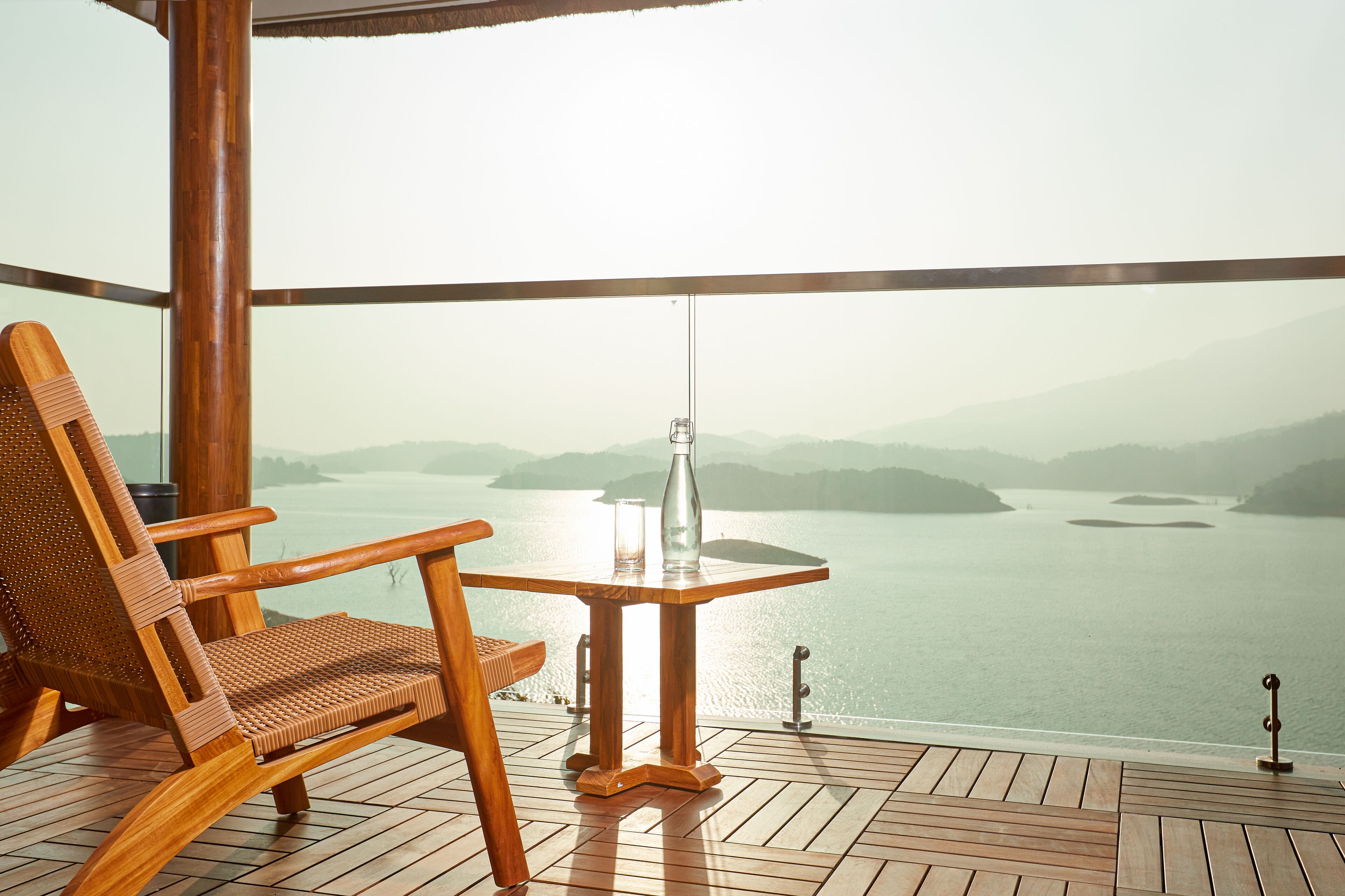 Une terrasse en bois avec une chaise en osier et une petite table, d’où l’on peut admirer un plan d’eau paisible et des collines lointaines.