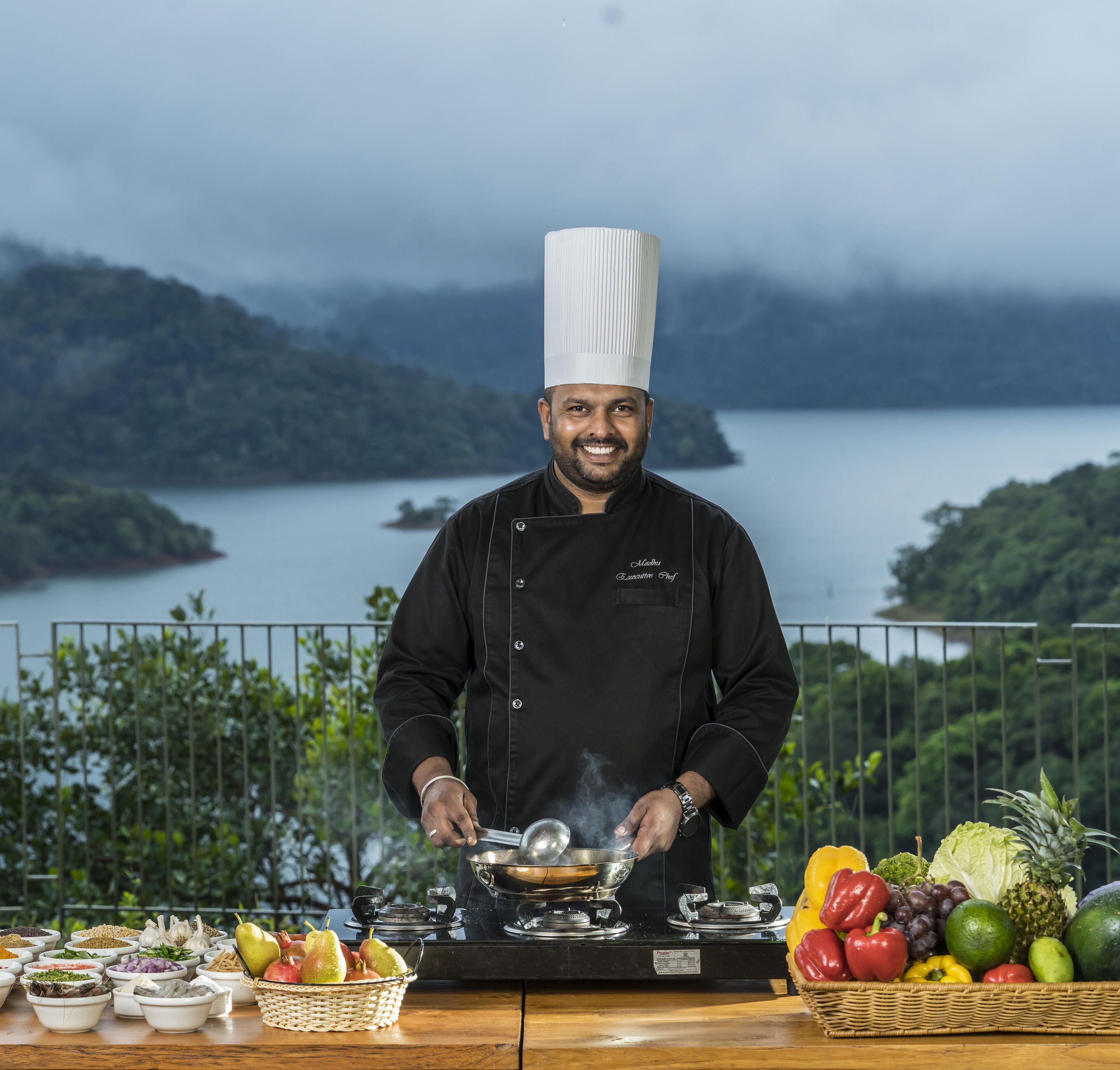 A chef cooking outdoors with a scenic view of a lake and mountains in the background.