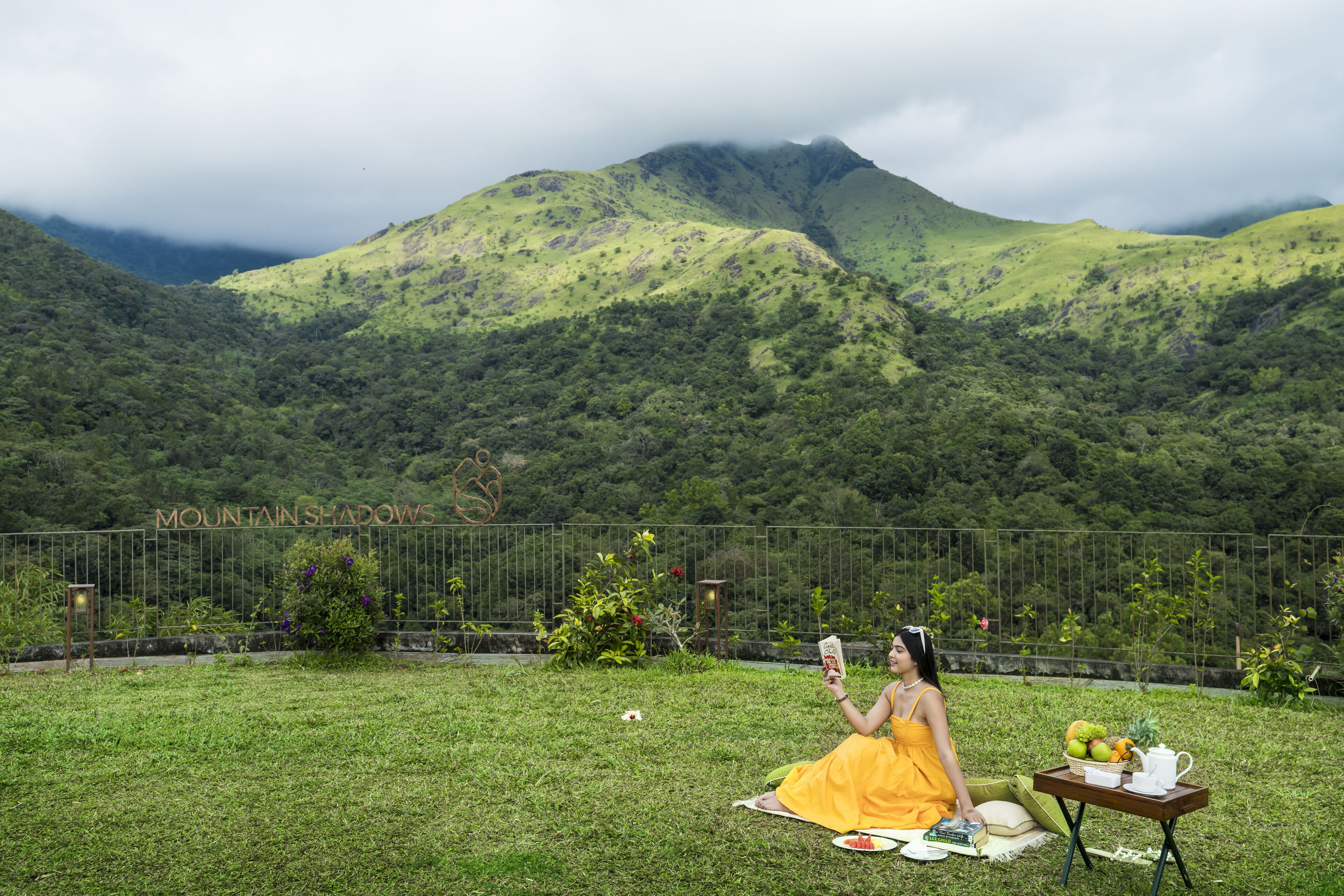 A woman in a yellow dress sitting on a blanket in a grassy area with a table of food and a book, with a mountainous landscape in the background.