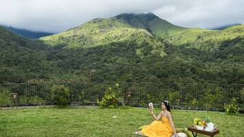 A woman in a yellow dress sitting on a blanket in a grassy area with a table of food and a book, with a mountainous landscape in the background.