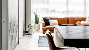 A modern living room with a brown leather sofa, a dining table with chairs, and large windows with blinds.