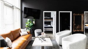 A modern living room with a brown leather sofa, a white coffee table, and a grey armchair. A flat-screen TV is mounted on the wall, and there is a large mirror with an ornate frame.
