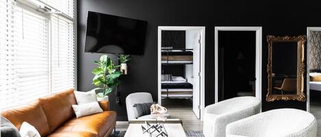 A modern living room with a brown leather sofa, a white coffee table, and a grey armchair. A flat-screen TV is mounted on the wall, and there is a large mirror with an ornate frame.
