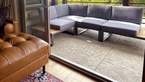 Uma sala de estar moderna com um sofá em formato de L, uma poltrona de couro marrom e uma porta de vidro que dá para uma varanda com vista para árvores e um edifício.