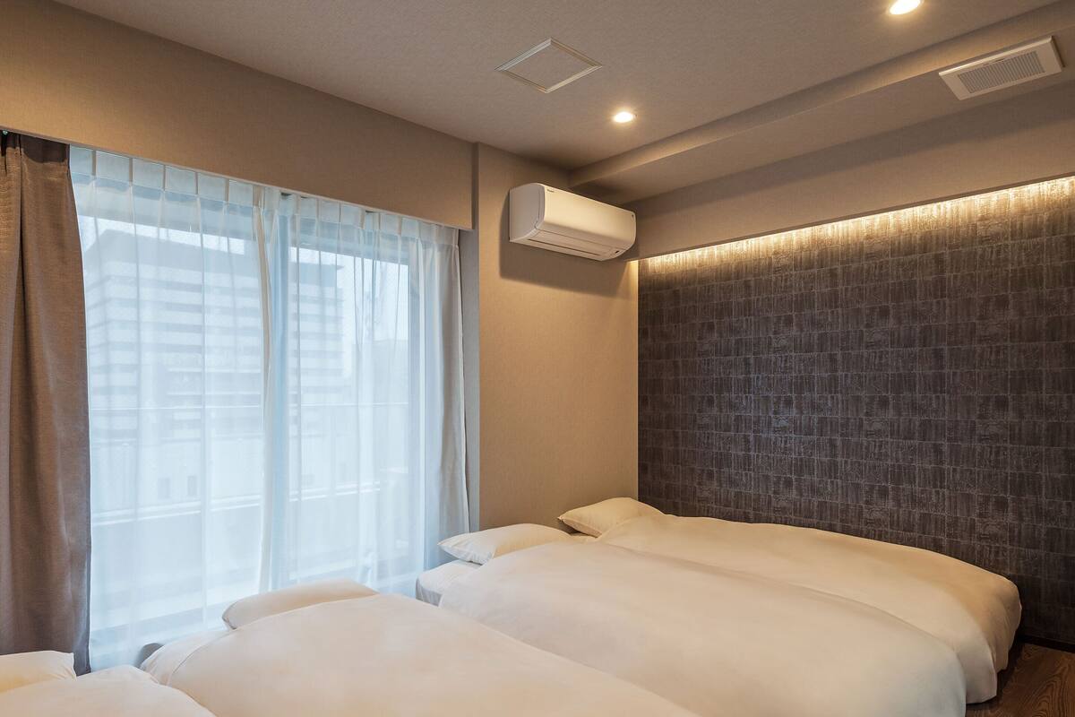 Habitación de hotel con dos camas, un ventanal con cortinas translúcidas, un equipo de aire acondicionado y una pared con textura.