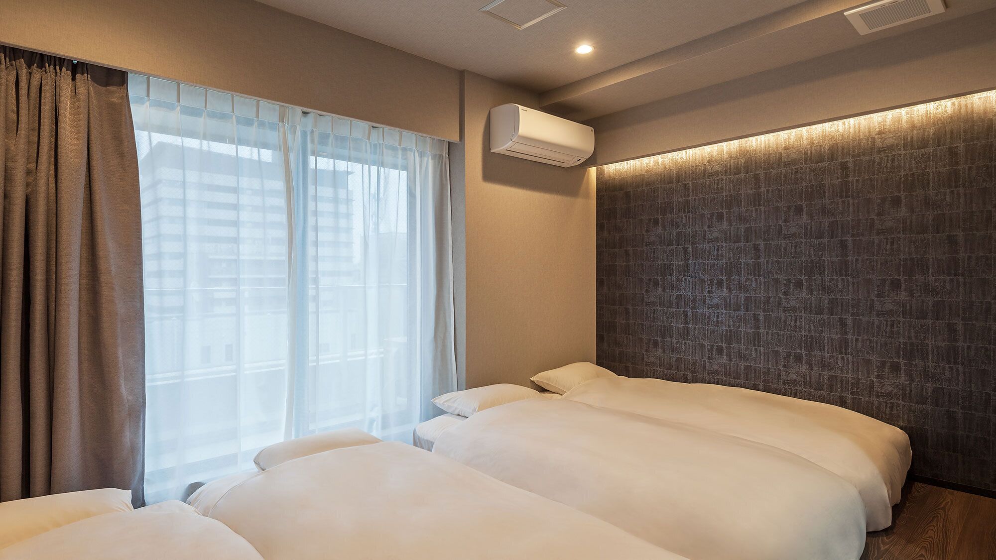 A hotel room with two beds, a window with sheer curtains, an air conditioning unit, and a textured wall.