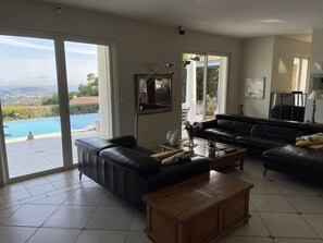 Ein geräumiges Wohnzimmer mit einer großen schwarzen Ledersofa, einem Holztisch und einem Fernseher. Ein großes Fenster bietet Blick auf einen Swimmingpool und Grünflächen.