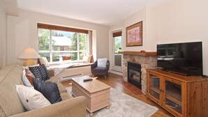 A living room with a sofa, armchair, coffee table, fireplace, TV, and large windows. - Eagle Lodge Village 2 bedroom condo, steps to Village, shops and restaurants (Whistler)