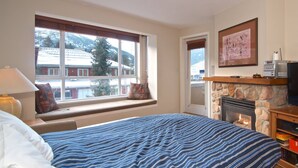 A bedroom with a large bed, a stone fireplace, a window with a view of buildings and trees, and a framed picture on the wall. - Eagle Lodge Village 2 bedroom condo, steps to Village, shops and restaurants (Whistler)