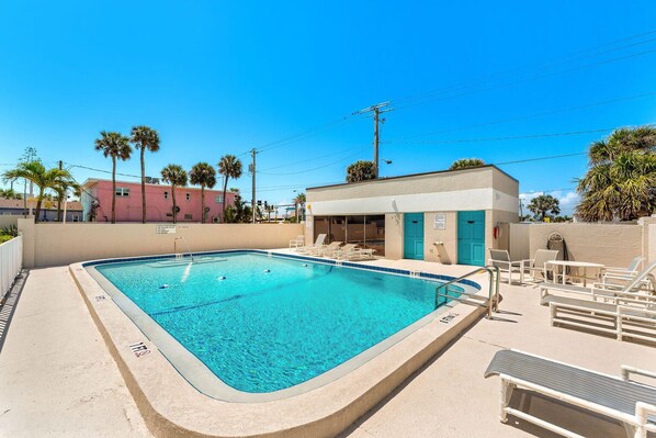 Una piscina rectangular con borde azul, rodeada de sillones de descanso y un pequeño edificio con puertas azules.