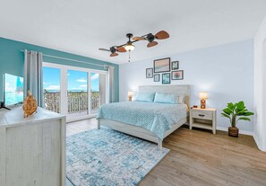 Un dormitorio con cama, mesitas de noche, ventilador de techo, ventana con vistas y puerta que da a un balcón.