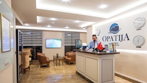 Área de recepção de hotel, com balcão, espaço de estar e parede exibindo relógios e o logotipo do hotel.