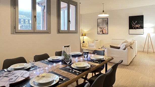 Un comedor con una mesa de madera preparada para cuatro personas, ventanas grandes, un sofá blanco y dos cuadros en la pared.