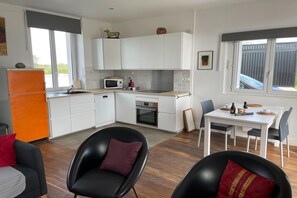 A compact kitchen with white cabinets, a microwave, and a sink. There is a dining area with a table and chairs, and a black leather chair with a red cushion.
