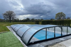 A swimming pool with a glass enclosure, surrounded by a paved area and a grassy field.