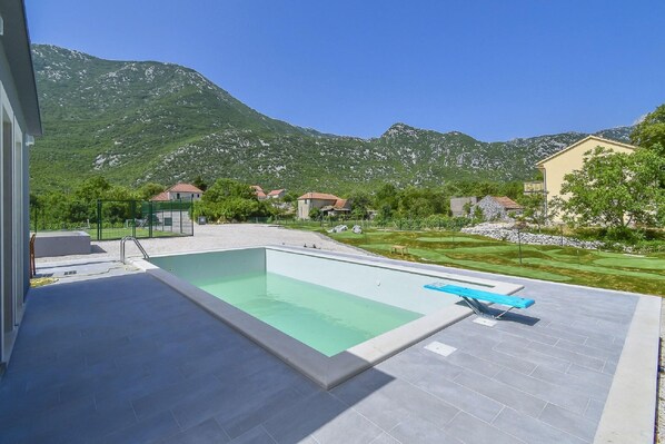 A swimming pool with a blue lounge chair, surrounded by a paved deck, with a mountainous backdrop and residential buildings.