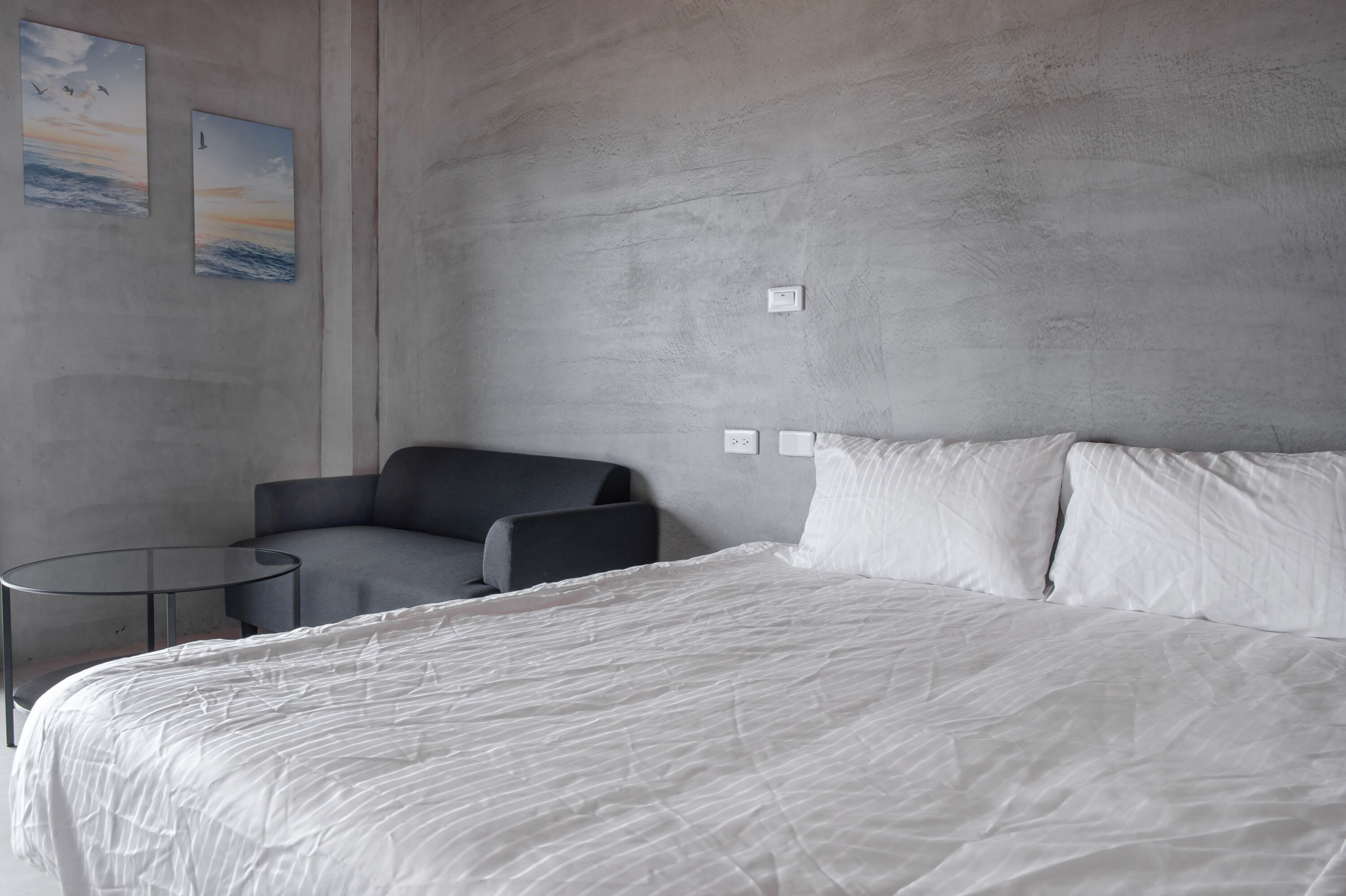 A modern hotel room with a large bed, a gray armchair, a small round table, and two wall-mounted light switches.