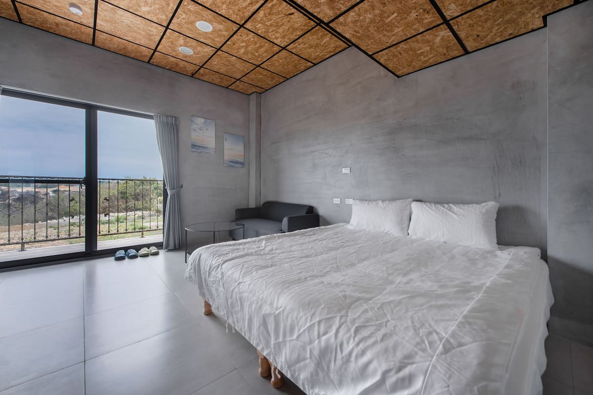A modern bedroom with a large bed, a small seating area, and a balcony with a view.