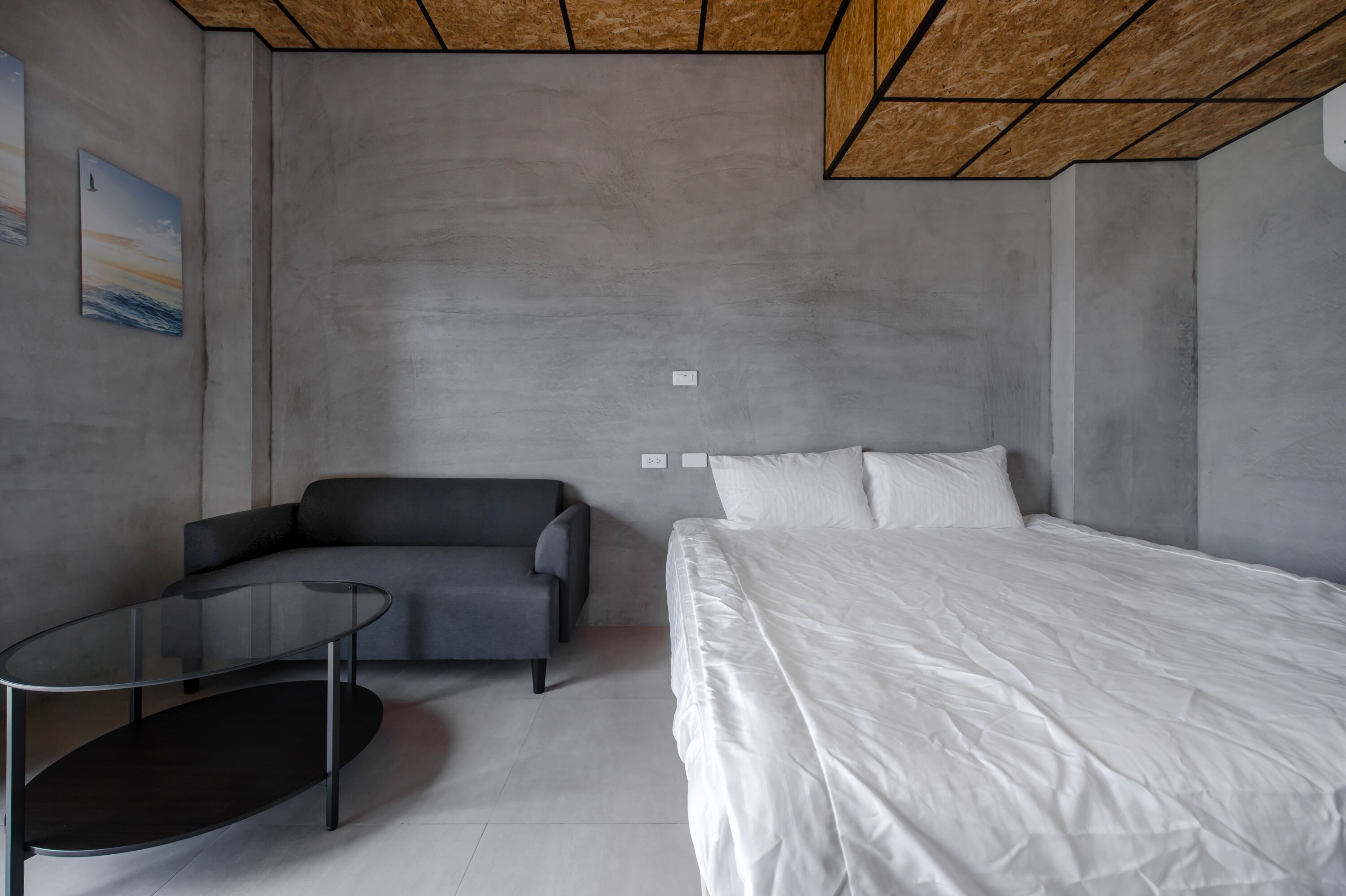 Un dormitorio moderno con una cama, una mesa de centro de vidrio, un sofá gris y un cuadro en la pared.