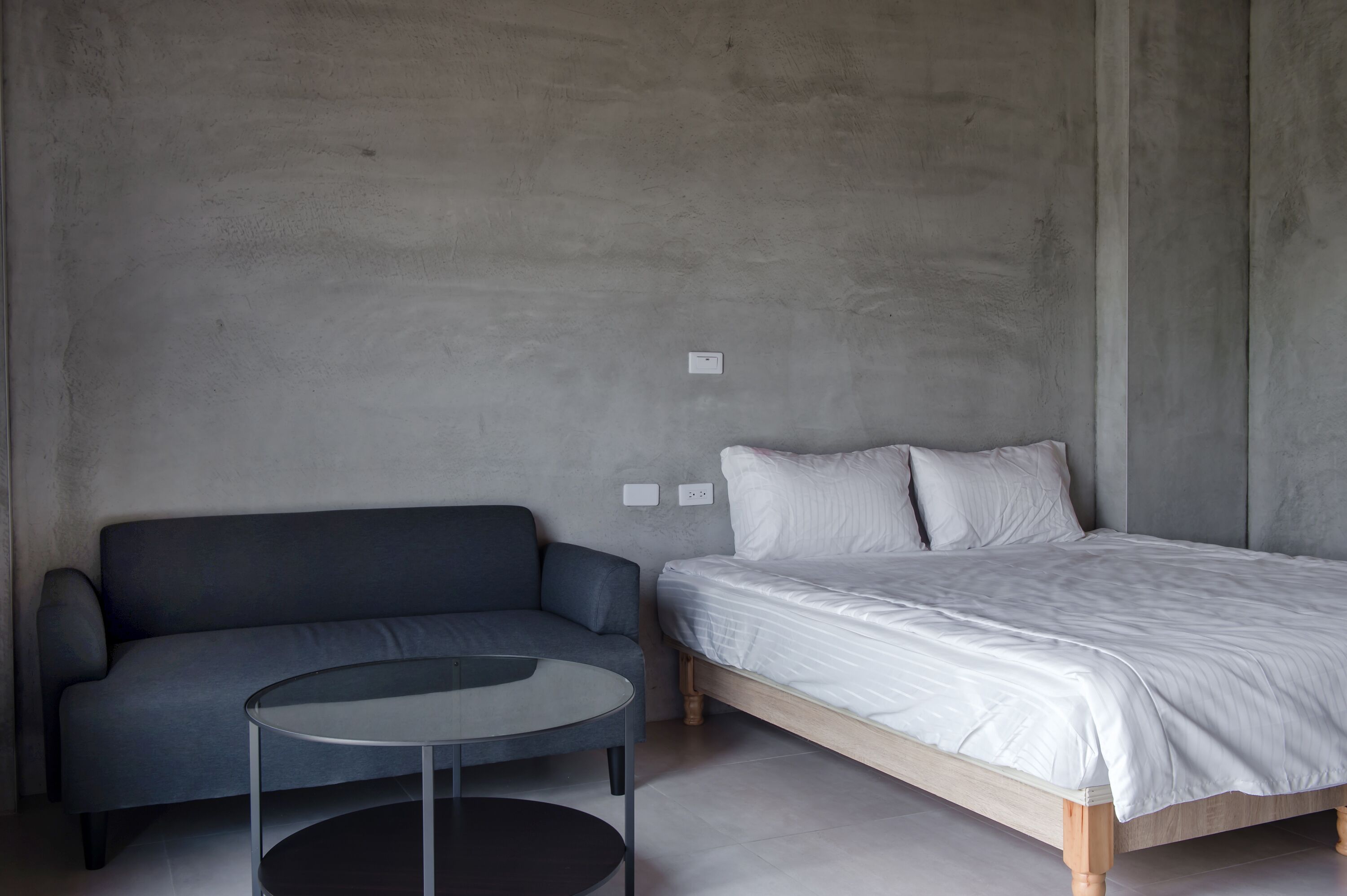 A minimalist room with a gray sofa, a round glass table, and a wooden bed with white bedding.