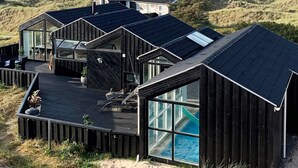 Un edificio moderno de madera con amplios ventanales y una terraza, con vistas a un paisaje costero de dunas y el mar.