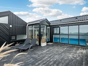 A modern outdoor deck with lounge chairs, a glass enclosure, and a view of the sea. - 4 Star Holiday Home in Saltum (Saltum)
