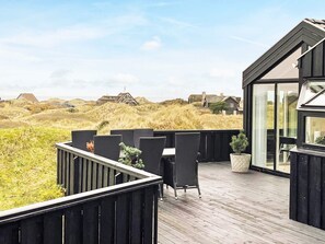 Um deck de madeira com móveis de exterior, com vista para uma paisagem de dunas e casas ao longe.