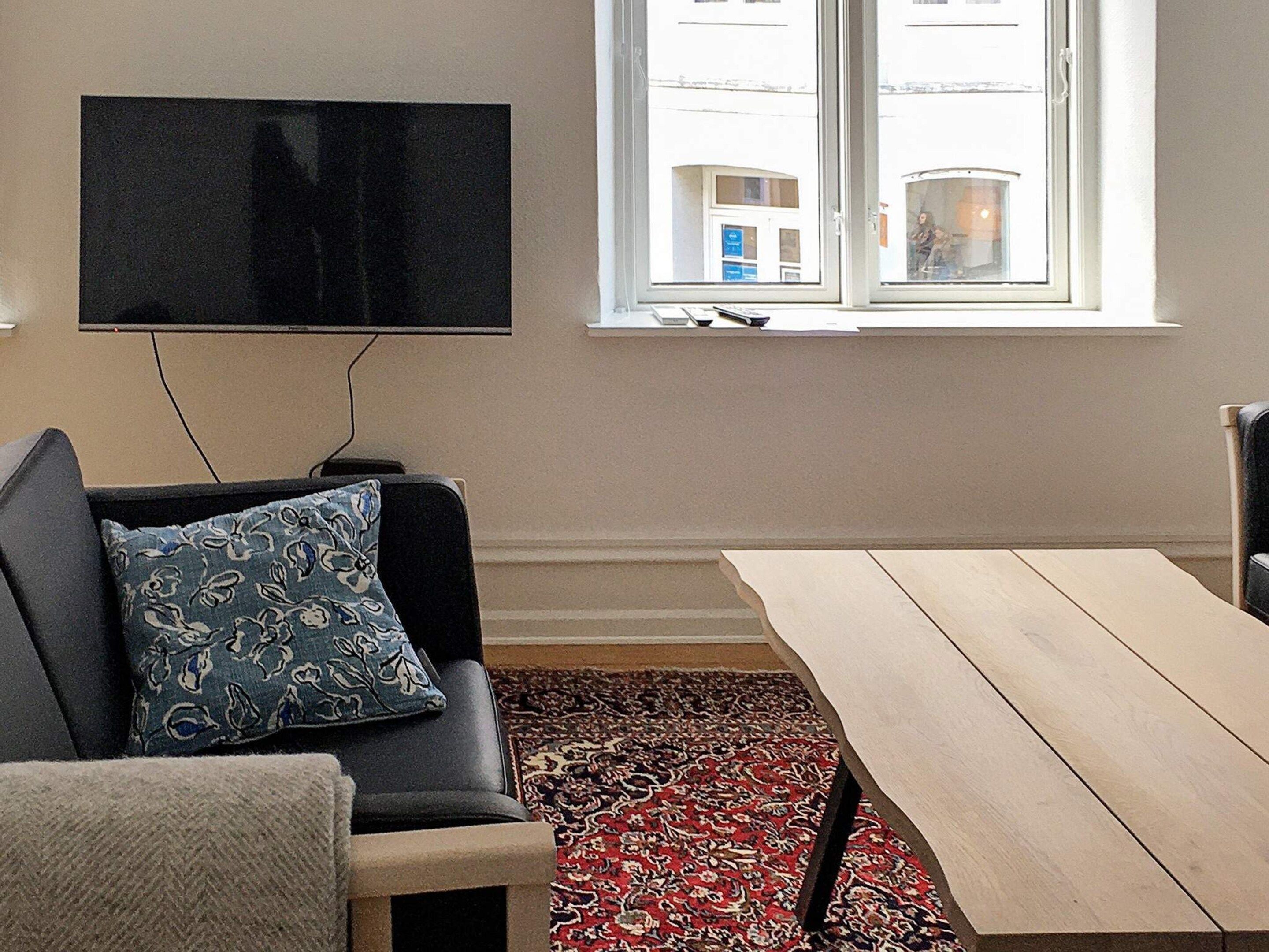 A living room with a sofa, a TV mounted on the wall, a window with a view of buildings, and a patterned rug.