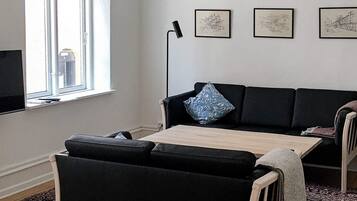 Un salon avec un canapé noir, une table basse en bois et un lampadaire. Trois œuvres d’art encadrées ornent le mur.