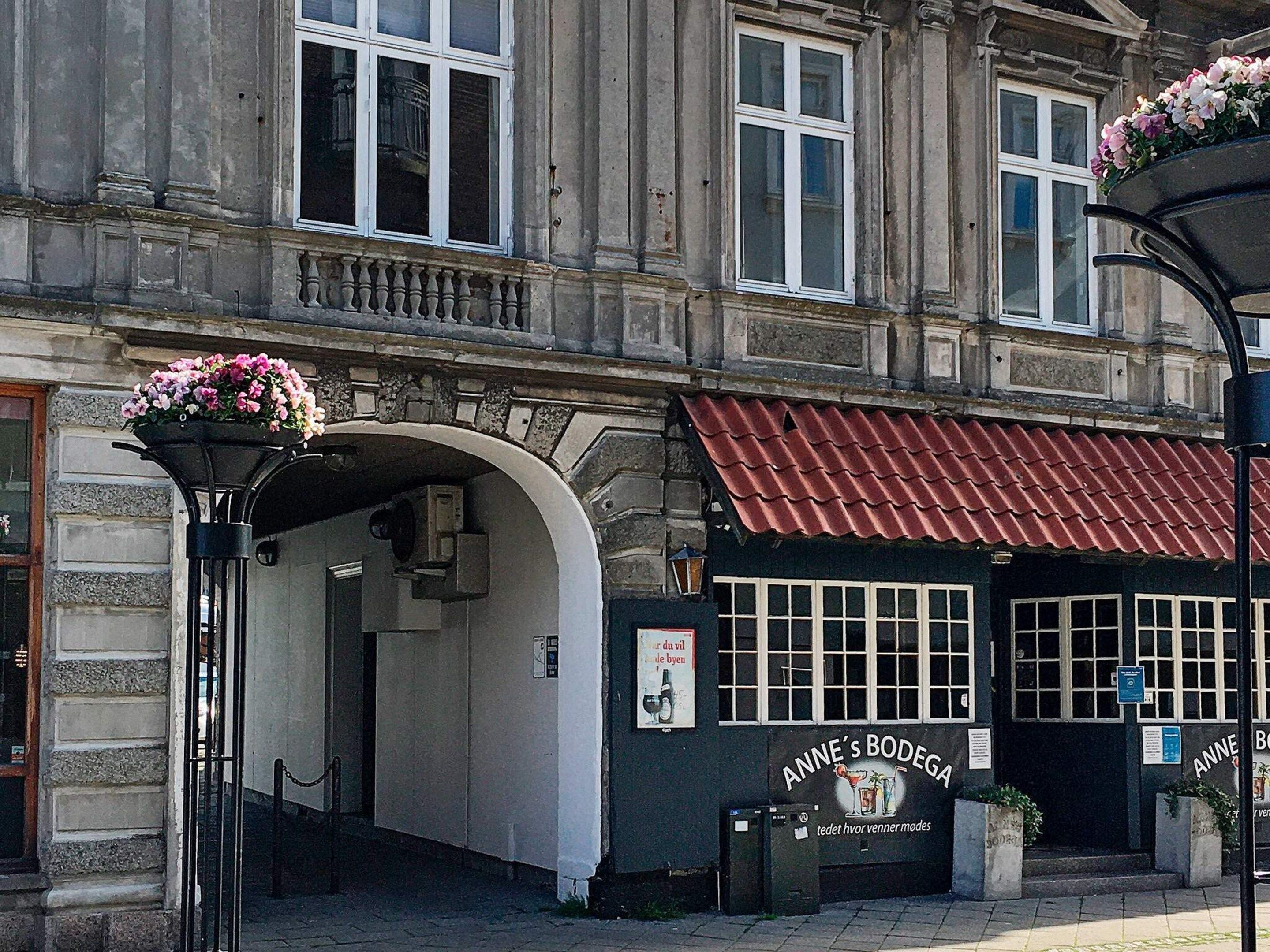 A building with a red-tiled roof, a sign reading 'Anne's Bodega,' and a flower basket on a post.