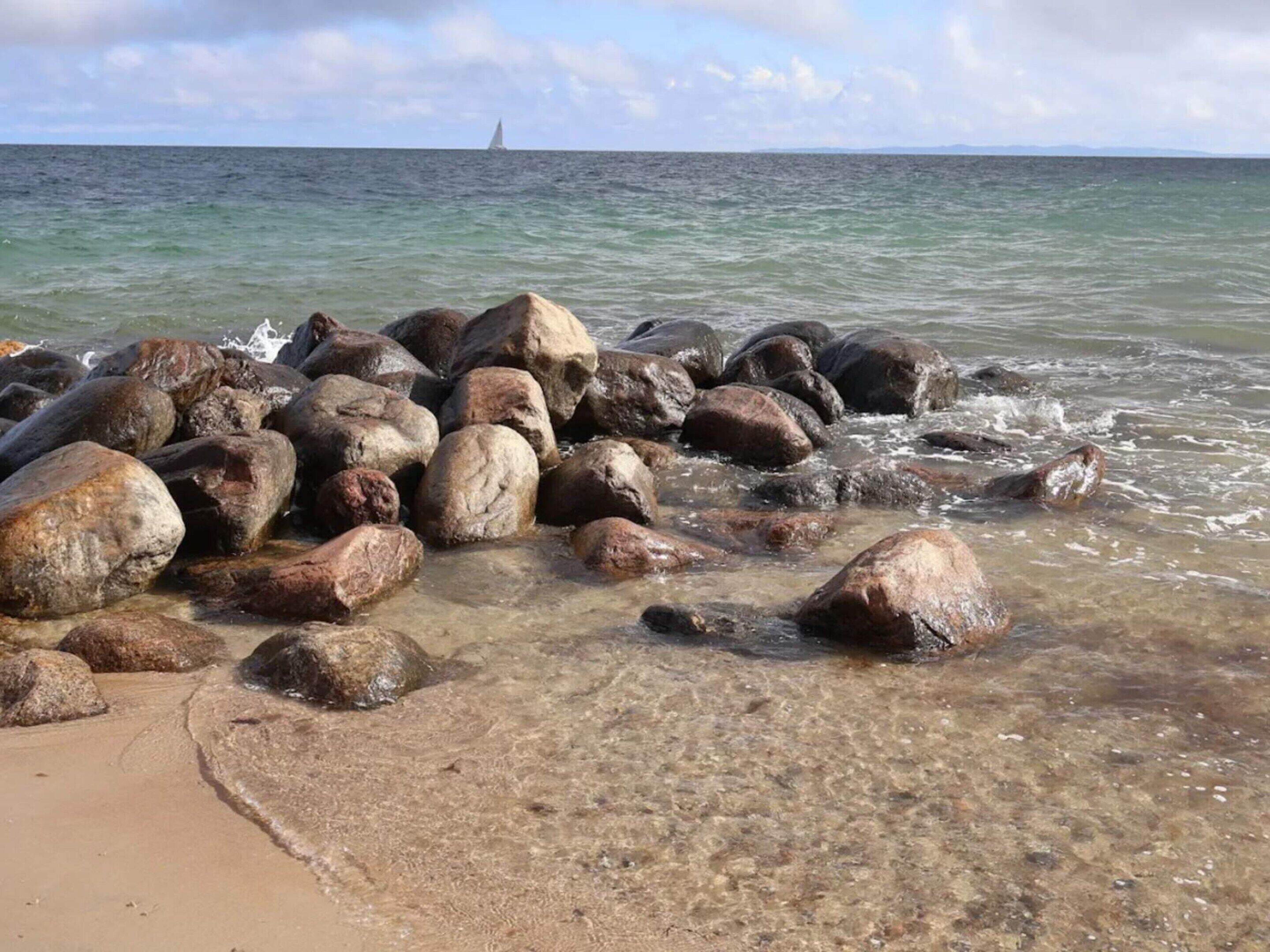 Una costa rocosa con agua cristalina y un velero a lo lejos.