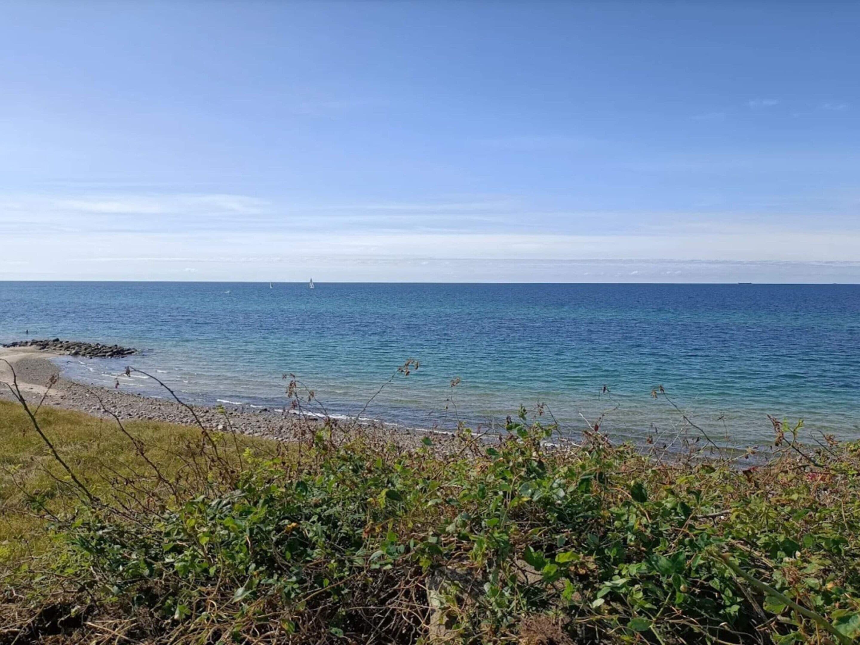 A coastal view with a sandy beach, clear blue sea, and distant sailboats.