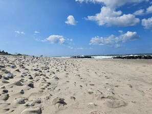 Una extensa playa arenosa con huellas y rocas, cielo despejado con nubes, y olas rompiendo contra la costa rocosa.