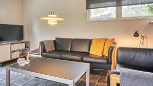 A modern living room with a black leather sofa, a grey armchair, a coffee table, a TV on a cabinet, and a window with blinds. - 4 Person Holiday Home in Aakirkeby (Akirkeby)