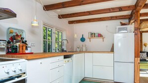Une cuisine compacte avec des meubles blancs, un plan de travail en bois, une cuisinière, un lave-vaisselle et un réfrigérateur. De grandes fenêtres laissent entrer la lumière naturelle et offrent une vue sur la verdure environnante.