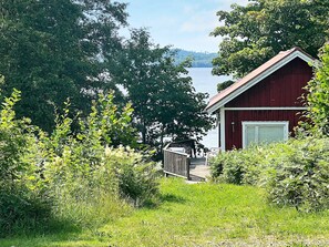 A red cabin with a deck overlooking a lake, surrounded by dense greenery. - 4 Person Holiday Home in Braas (Braås)