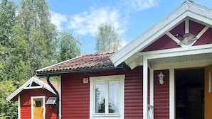 Een rode houten chalet met een veranda en een klein bijgebouwtje, een tafel met stoelen, en een deur die naar binnen leidt.