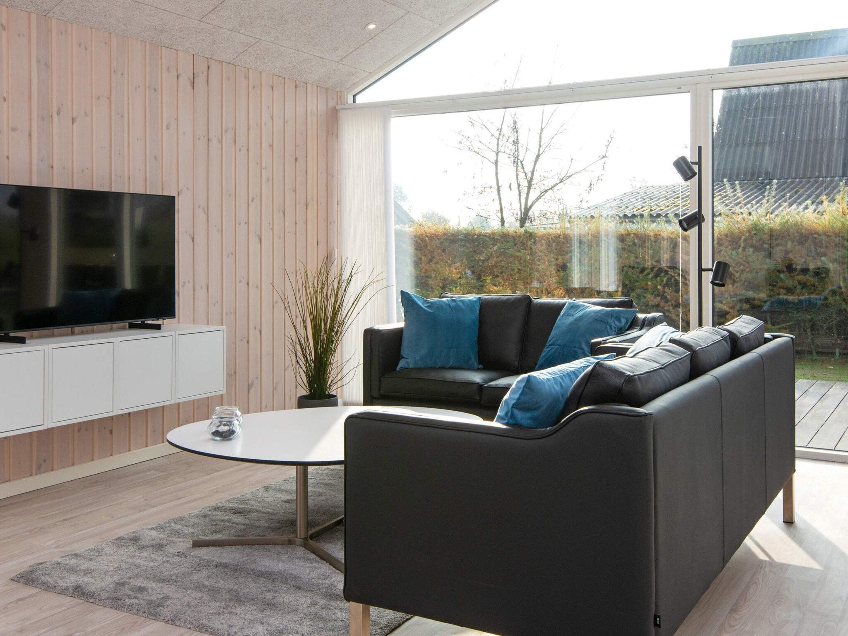 A modern living room with a flat-screen TV, a black sofa with blue cushions, a white coffee table, and a large window offering a view of the outdoors.