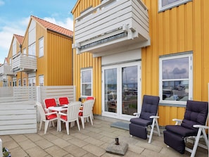 A yellow building with a patio area featuring a table and chairs, a sliding glass door, and a view of boats. - 4 Person Holiday Home in Rudkobing (Rudkobing)
