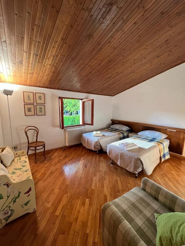 Een knus zolderkamer met houten vloeren, twee eenpersoonsbedden, een bank, een stoel en een raam met uitzicht op groen.