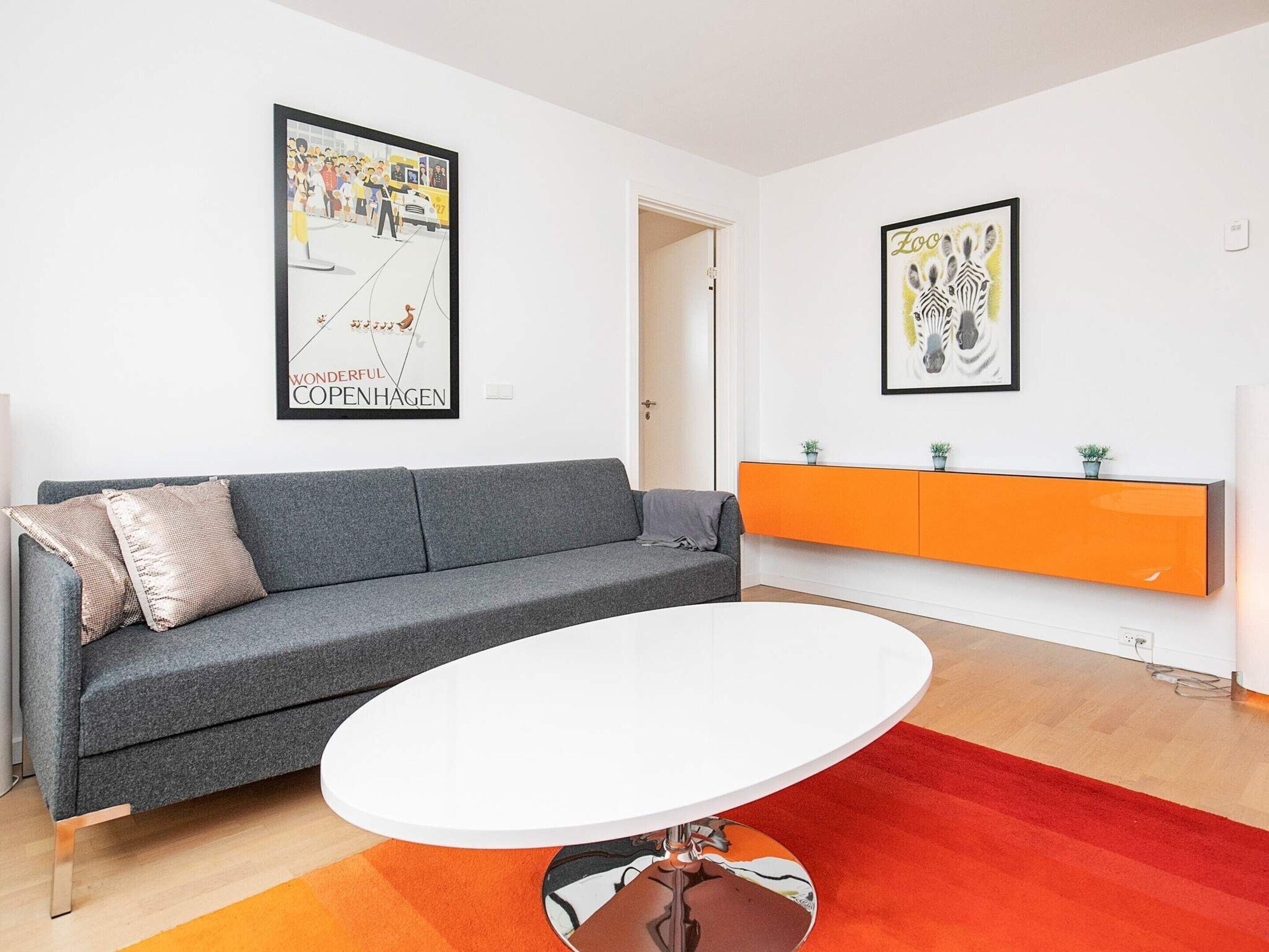 A modern living room with a grey sofa, a white coffee table, and an orange cabinet. There are two framed posters on the walls.